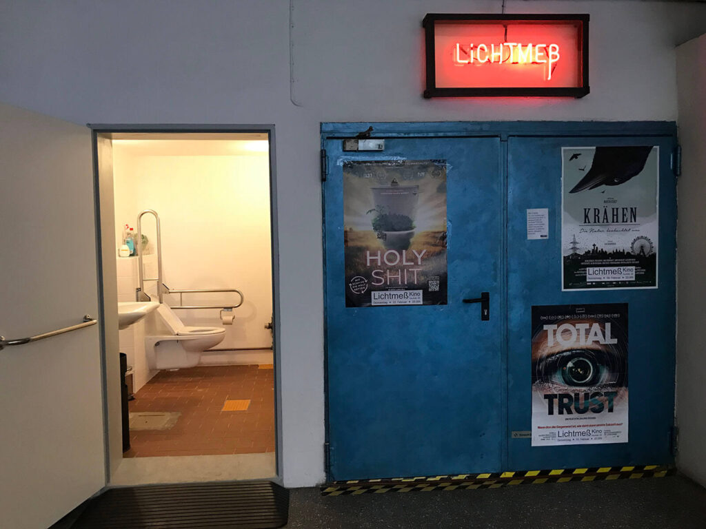 Ein Foto von einer Stahltür, dem Eingang zum Kinosaal, links daneben eine offene Tür zu einem Raum mit einer barrierefreien Toilette